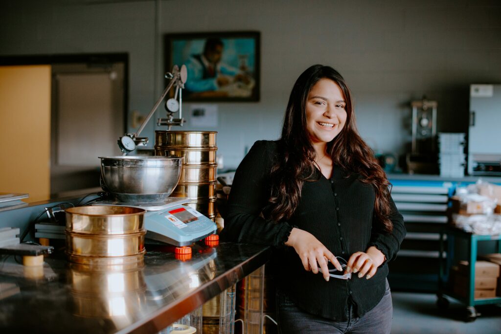 A student in a black shirt stands smiling next to scientific equipment on a laboratory counter.