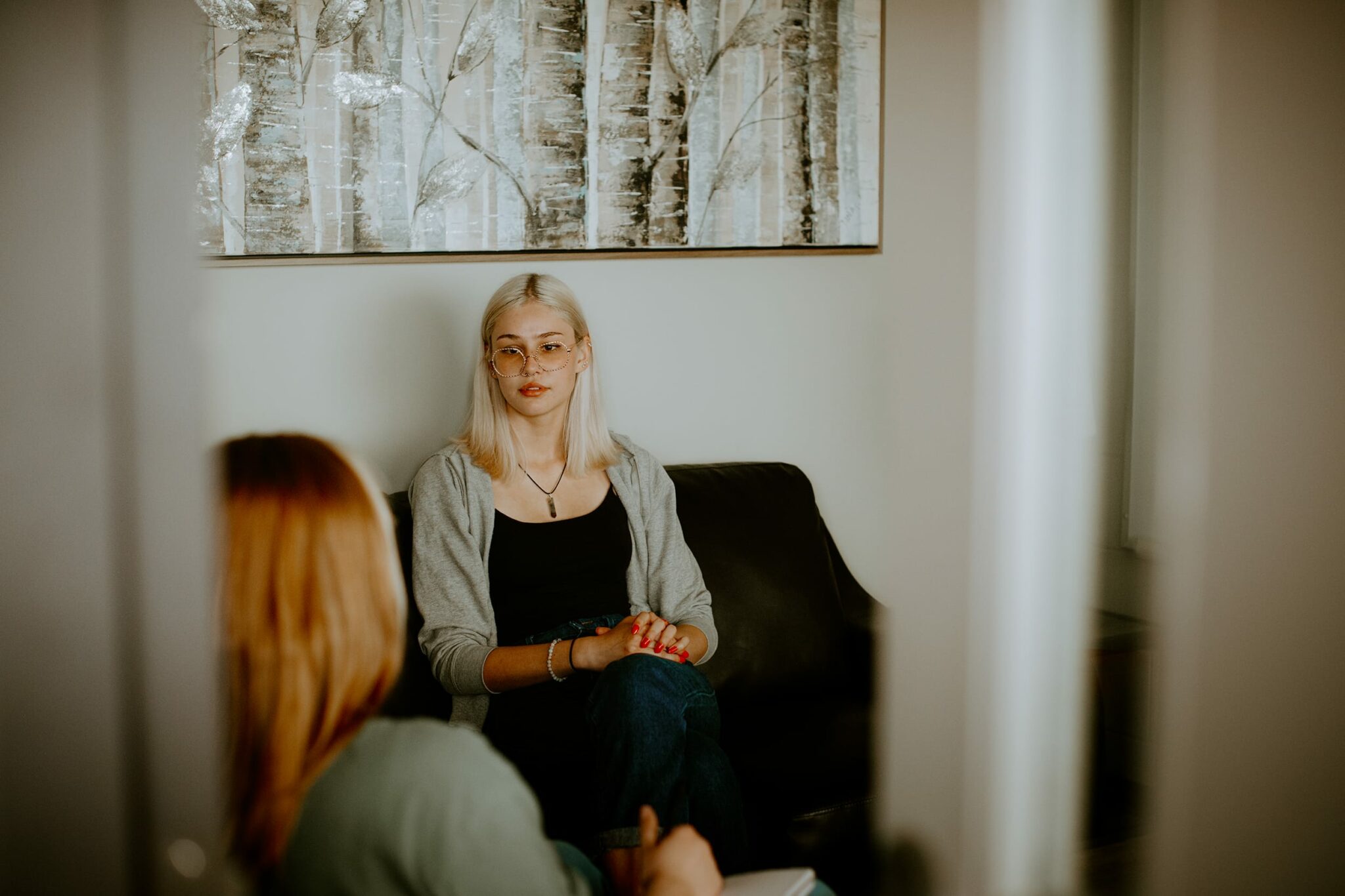 Two individuals seated in a room having a conversation, with one facing the camera and the other seen from behind. A forest-themed painting is on the wall behind them.