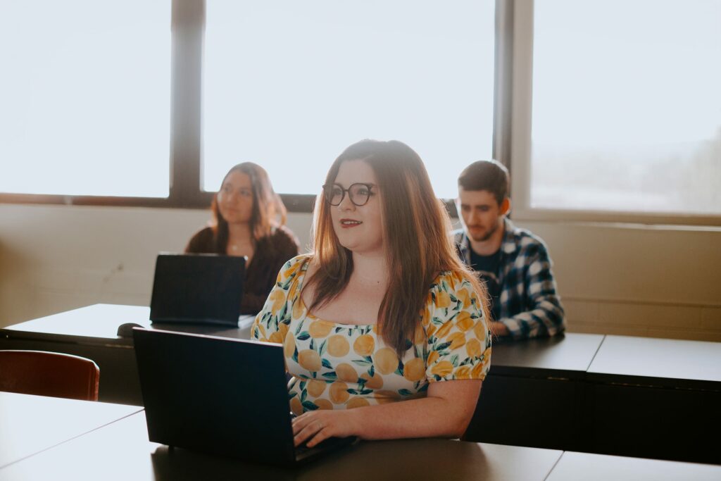 Three students sitting at desks with laptops in a classroom, with bright sunlight coming through large windows.