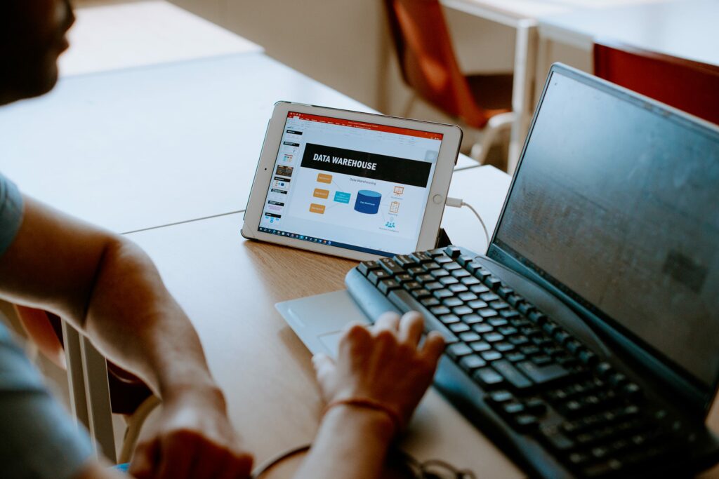 A student working at a desk with a laptop and a tablet displaying a data warehouse presentation.