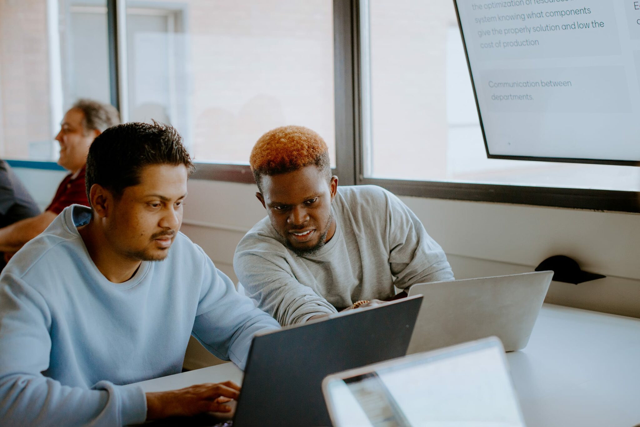 Two students collaborate at a table with laptops. A whiteboard in the background displays text.