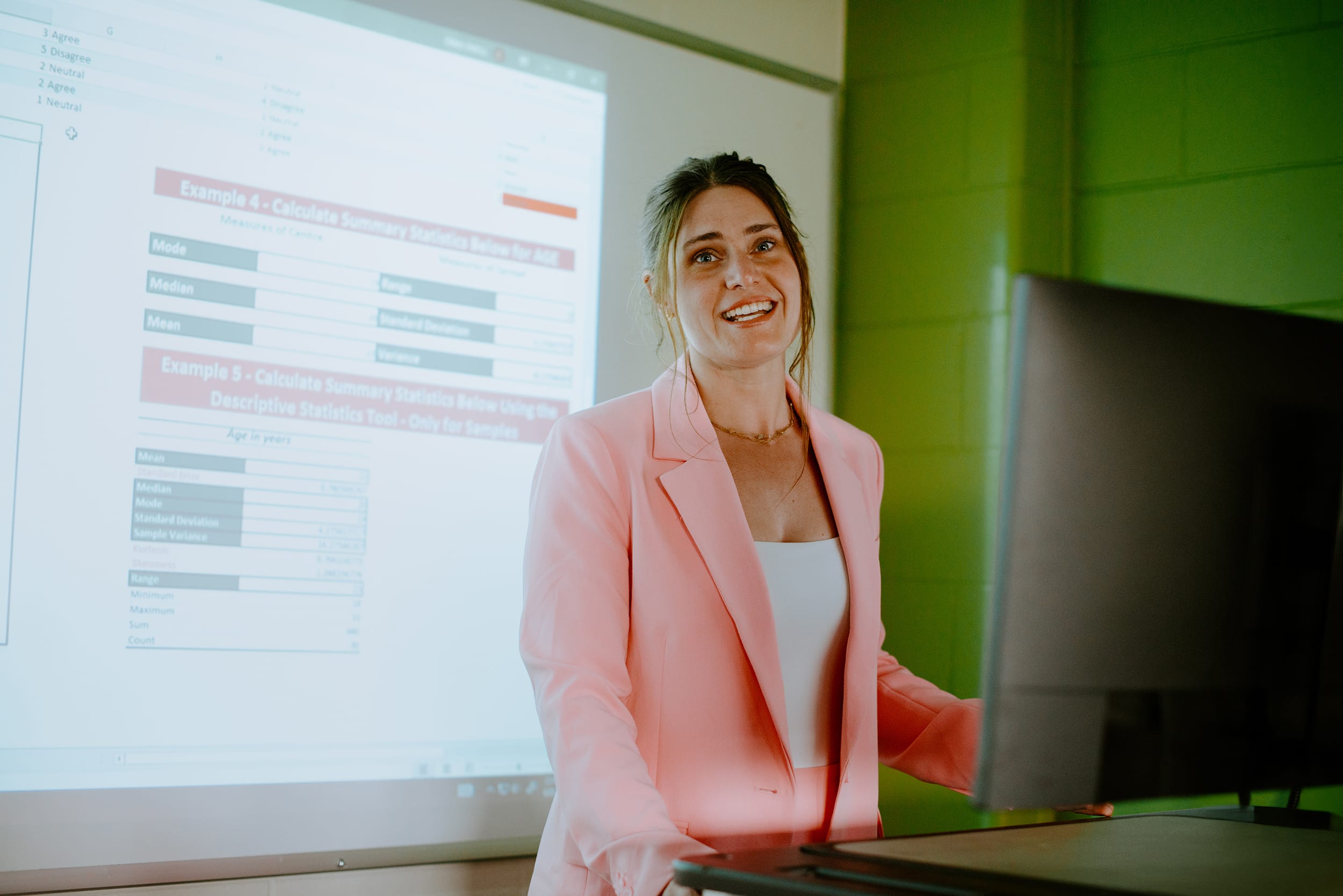 An individual in a pink blazer presents data on a screen using a computer.