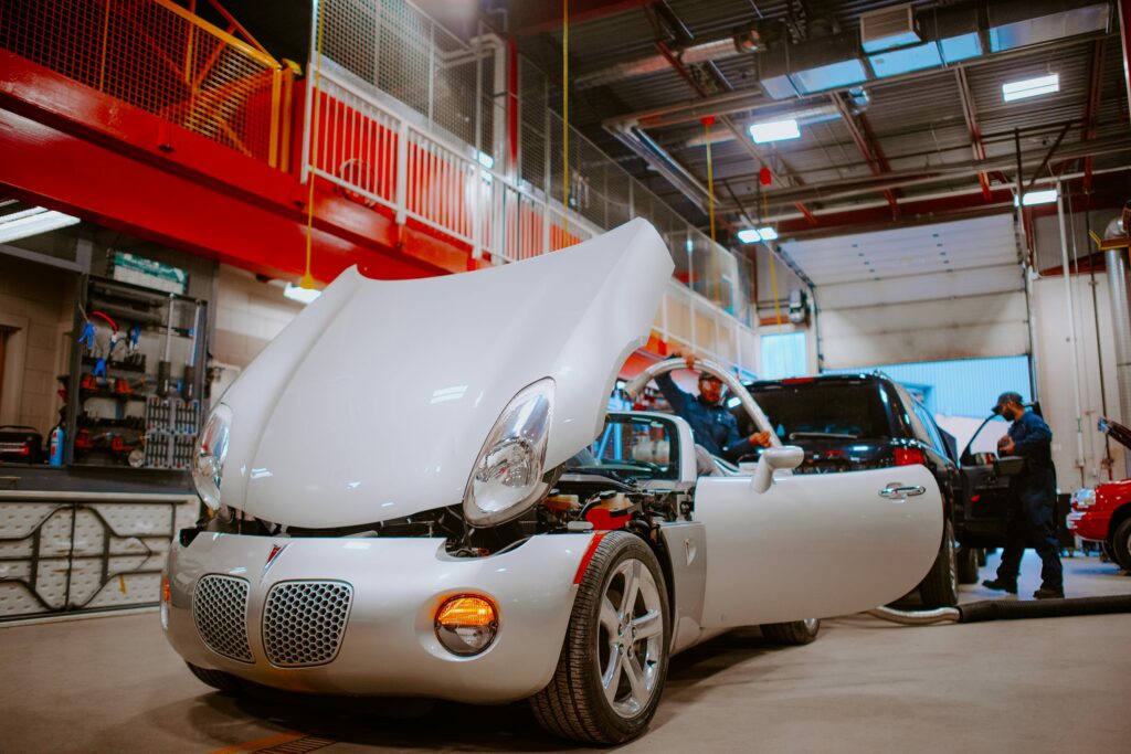 A white Pontiac Solstice with its hood open is being serviced in a garage. Mechanics are working on it, with various tools and equipment visible around the vehicle.