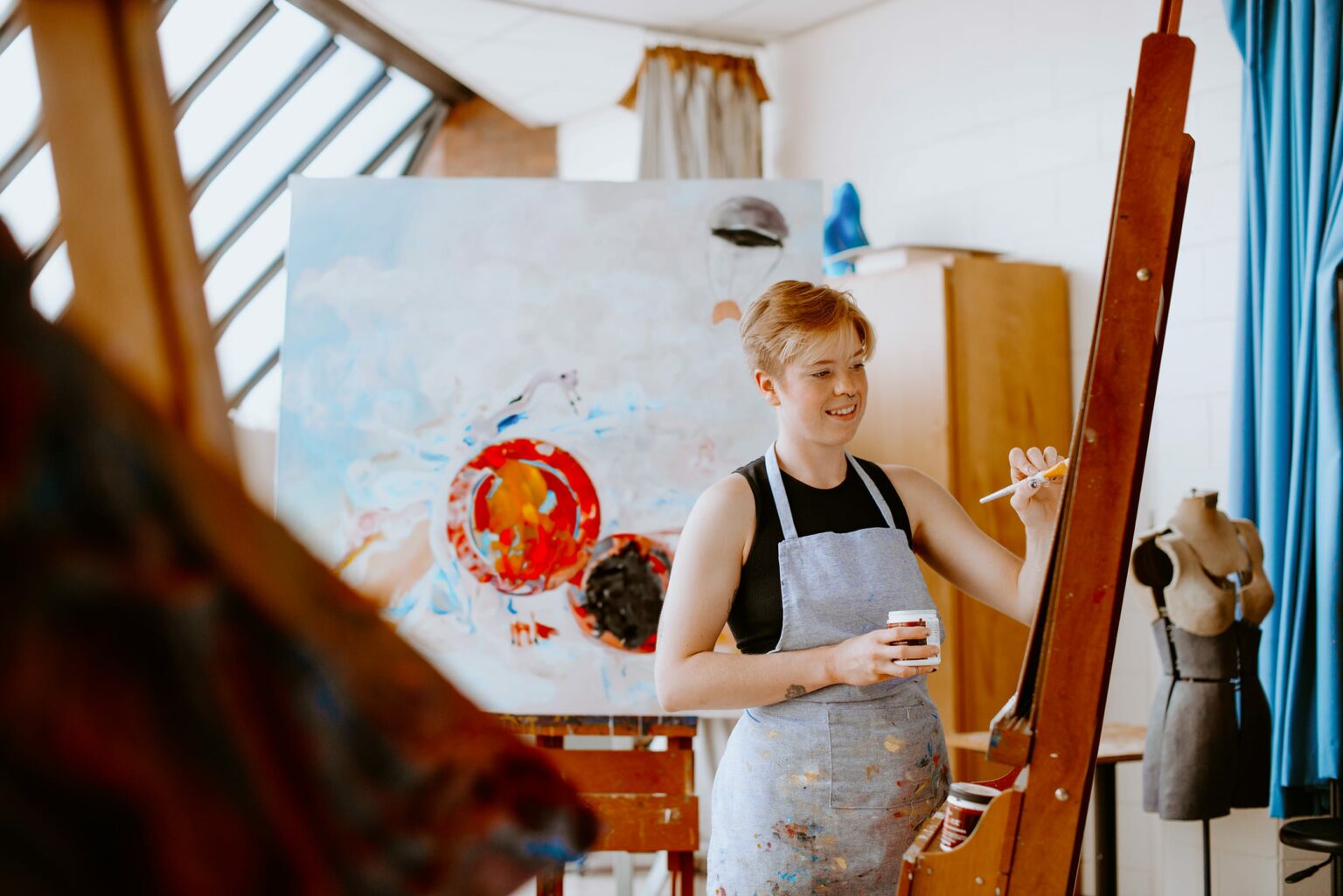 Artist painting on canvas in a studio with a mannequin nearby.