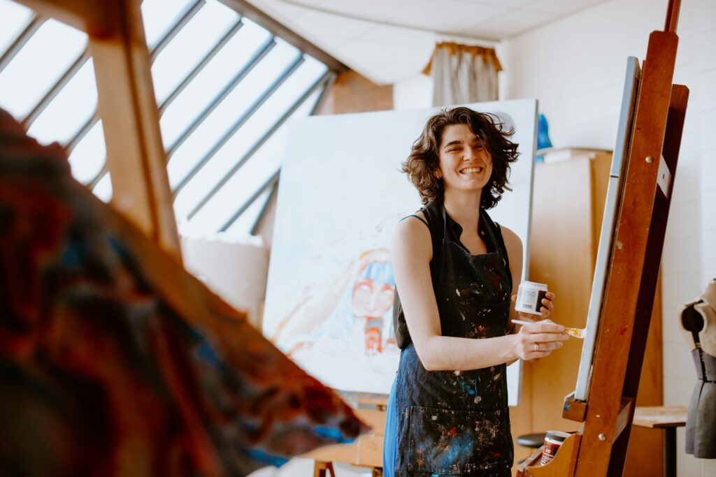 Student smiling while painting on a canvas in a bright studio. They are holding a paintbrush and a container of paint, wearing a dark apron.