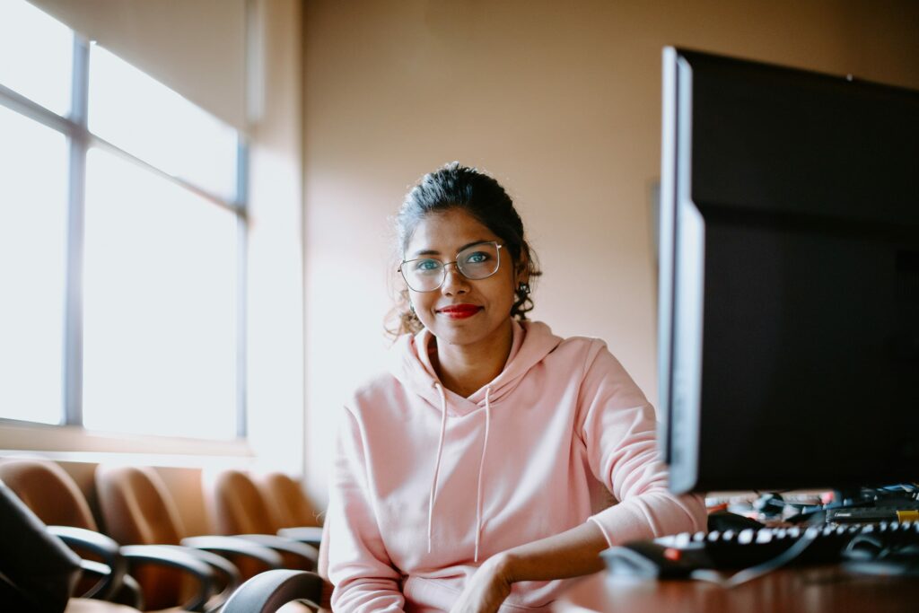 A student wearing glasses and a pink hoodie sits at a desk in front of a computer in a well-lit room.