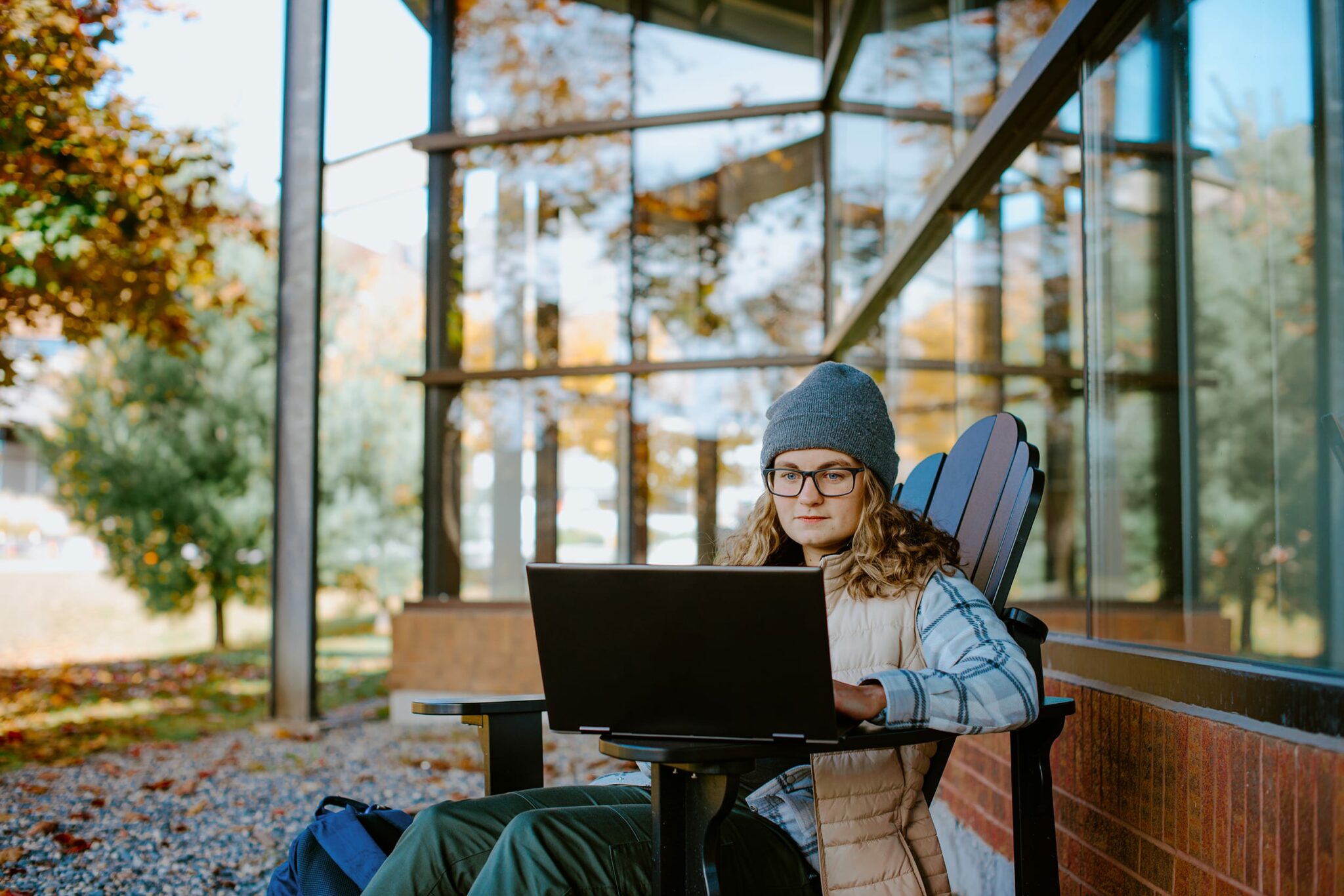 A student wearing a beanie and glasses works on a laptop while seated outside a building with large windows.