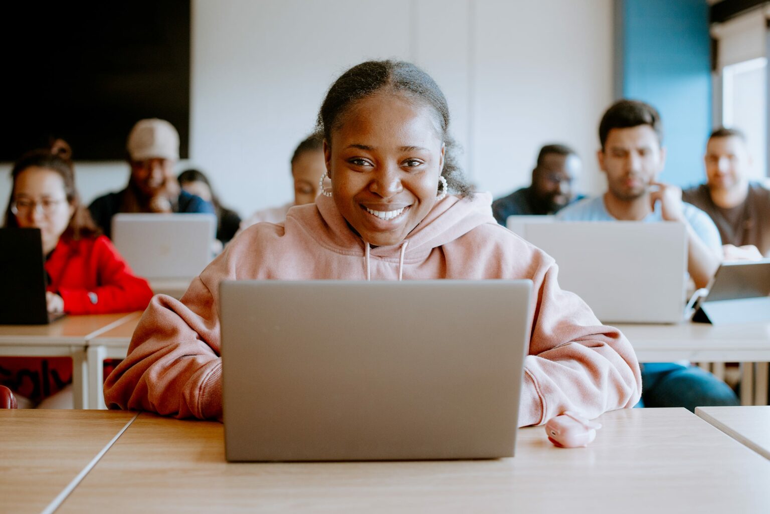 Student in a pink hoodie sits at a desk using a laptop, smiling. A classroom setting with others working on laptops in the background.
