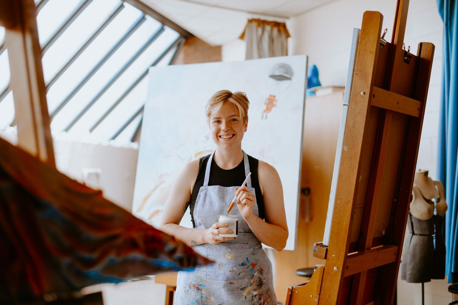 Student in an apron stands smiling, holding a paintbrush and cup in an art studio. Artwork and an easel are in the background.