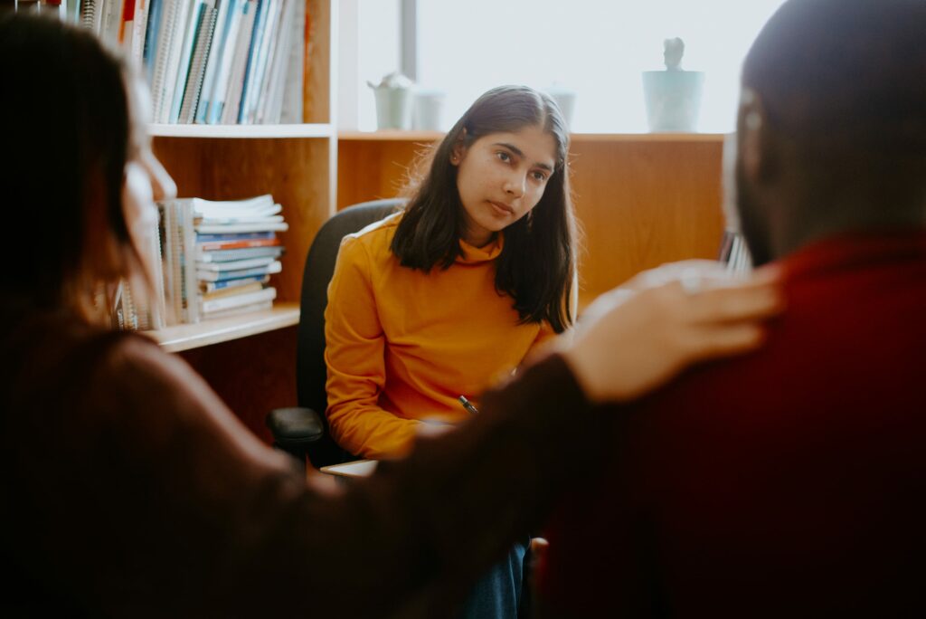 Three students sit in an office, engaged in a conversation. A student in a yellow shirt sits opposite, listening attentively, while another student has a hand on another's shoulder. Bookshelves are visible.