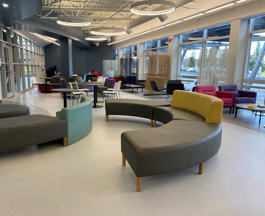 Cambrian's Student Life Centre Fireside Lounge. A brightly lit modern lounge with colorful curved seating, tables, and chairs. Large windows line the right side, and circular lights hang from the ceiling.