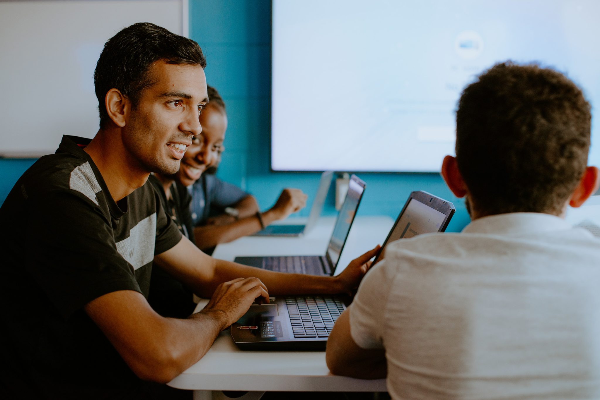 3 students sitting in a computer lab with their laptops and talking to each other.