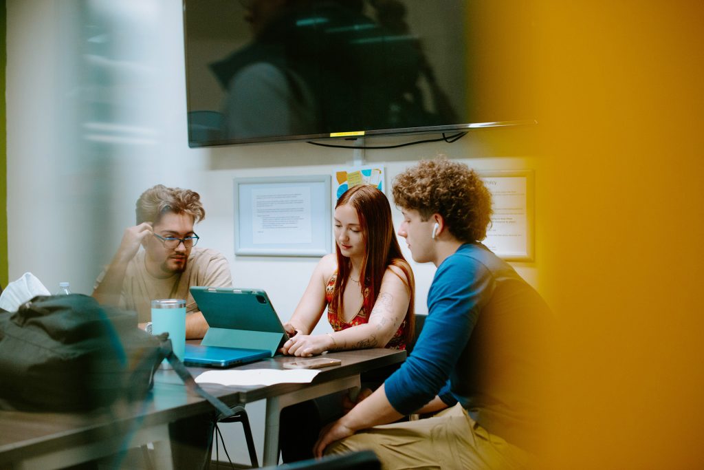 Three students sit around a table with a laptop in a meeting room, engaging in discussion.