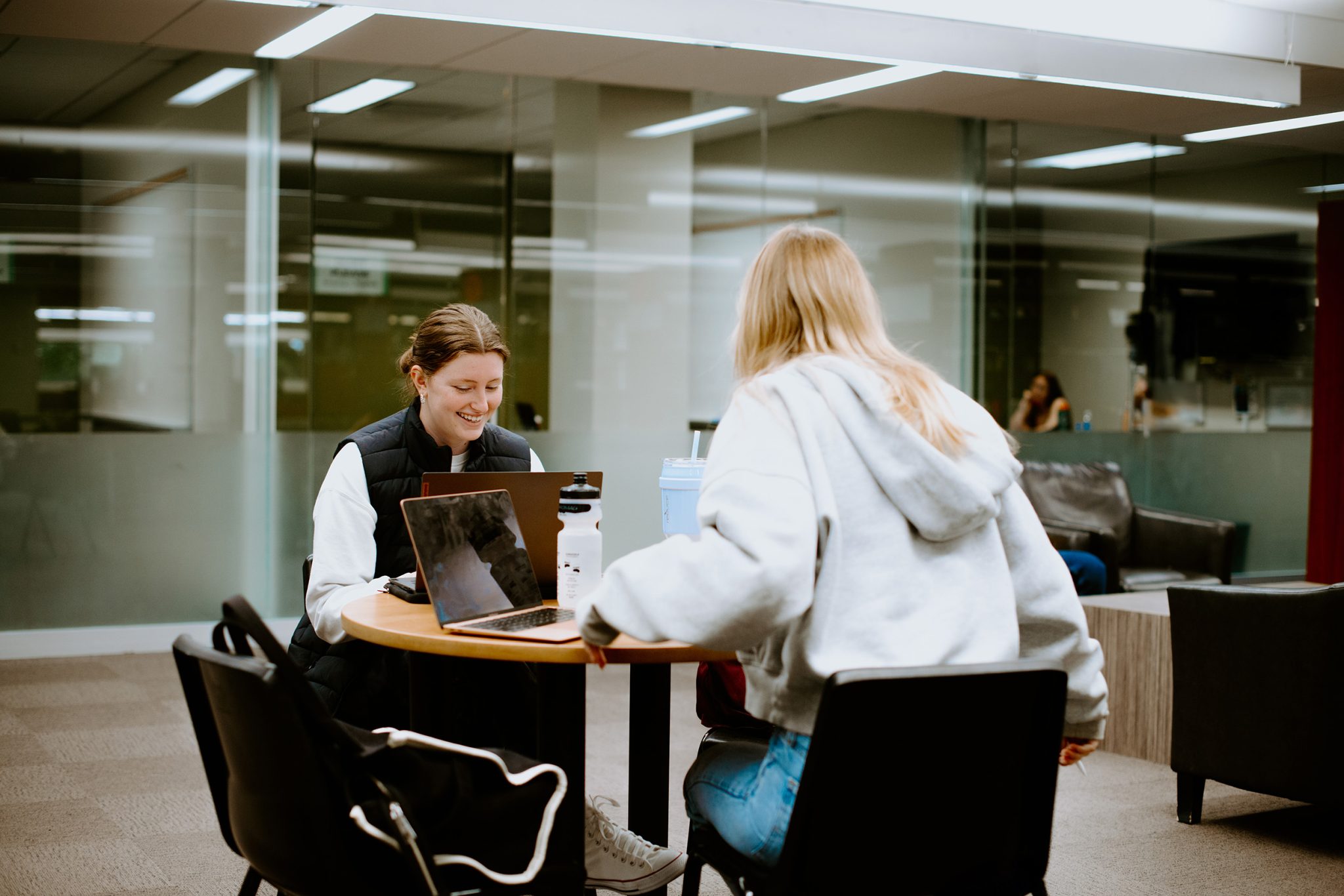Two students sit at a round table in a modern indoor setting, both using laptops. One person faces the camera, smiling. Chairs and a glass wall are visible in the background.