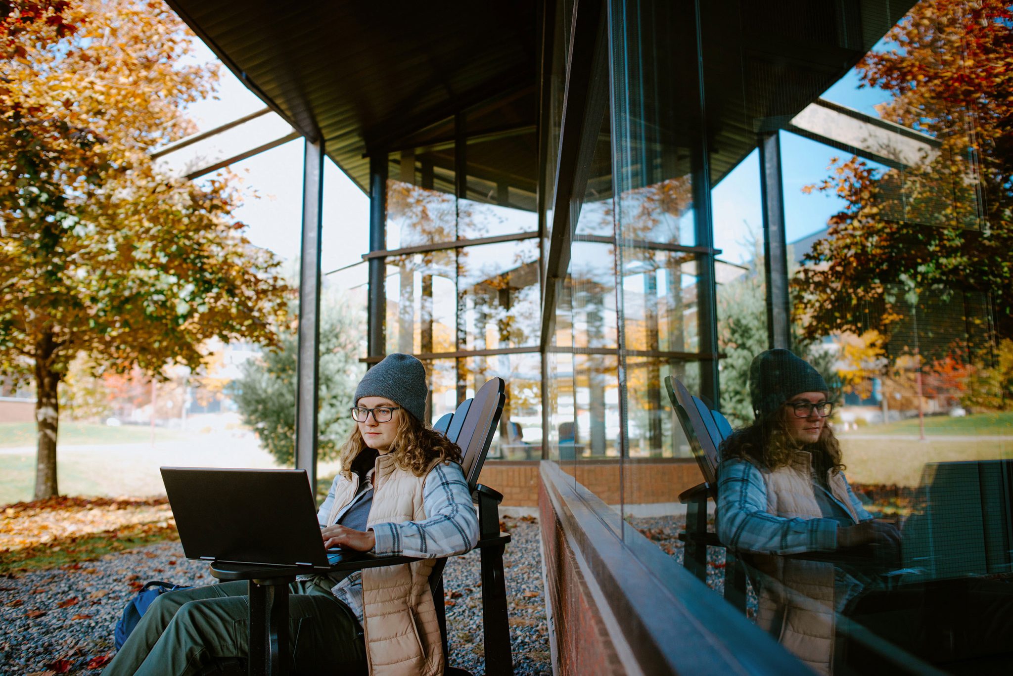 Student sitting on a chair outdoors using a laptop near a reflective glass building, surrounded by autumn trees.