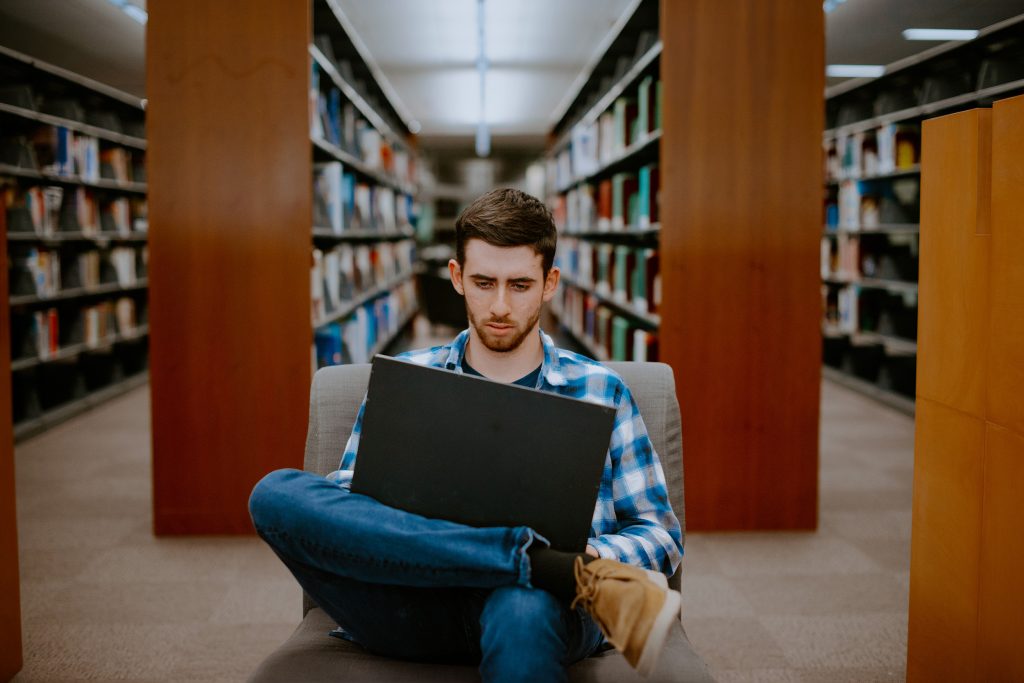 A student in a blue plaid shirt and jeans sits on a chair, concentrating on a laptop in a library aisle lined with books.