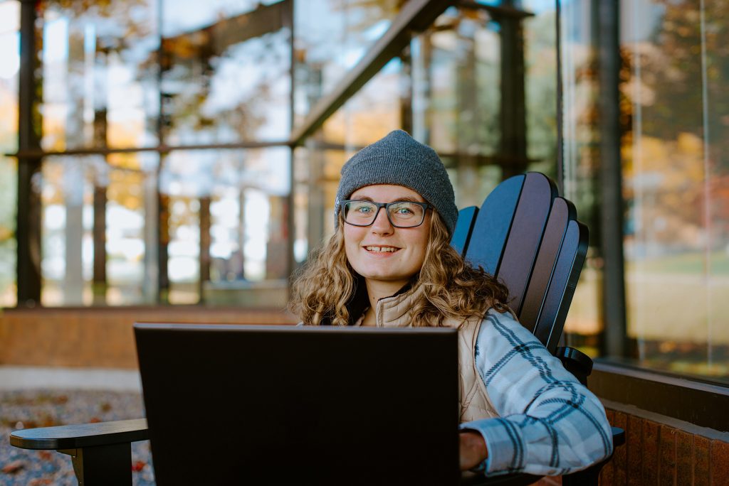 A student wearing a beanie and glasses sits on a wooden chair outdoors, using a laptop. The background shows a glass building with autumn trees reflected in it.