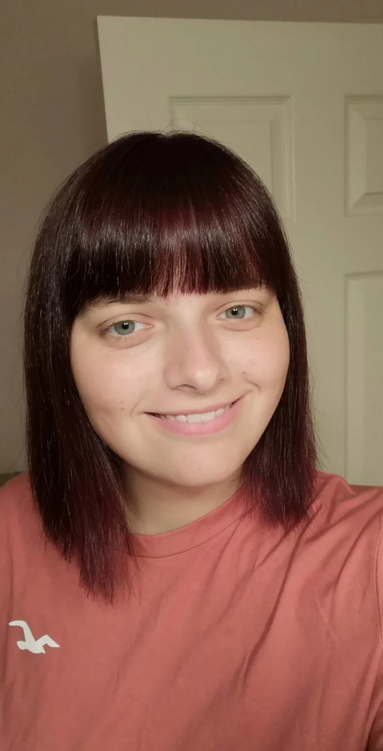 A person with shoulder-length dark hair and straight bangs, wearing a salmon-colored shirt, stands smiling in front of a light-colored door, perhaps an Analytics student from Cambrian College enthused by learning about various organizations.