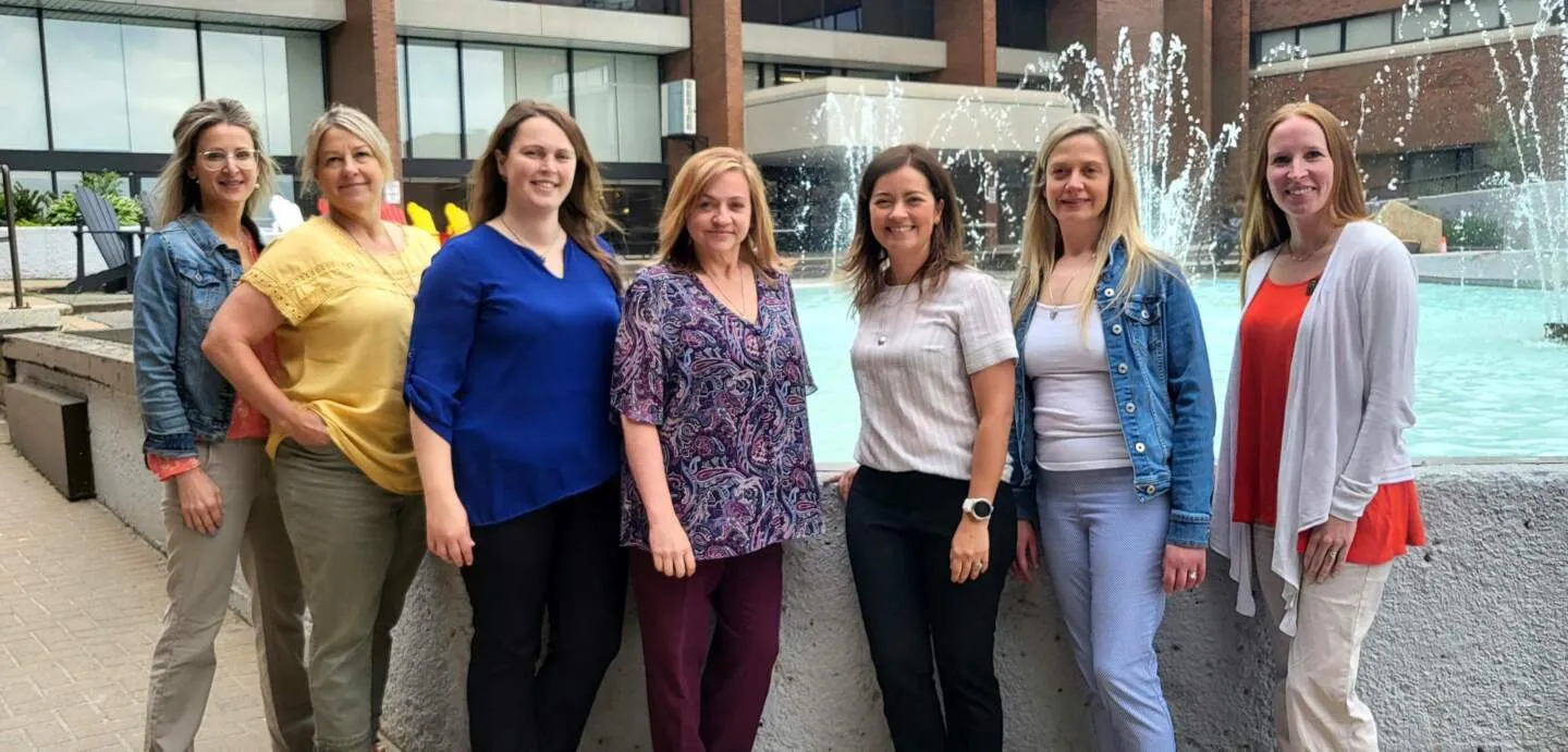 Seven women, including students from Cambrian College, stand in front of a fountain and building, smiling at the camera.