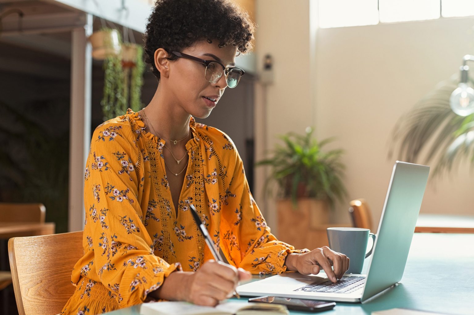Woman at desk in yellow shirt working on a laptop and writing in a notebook.