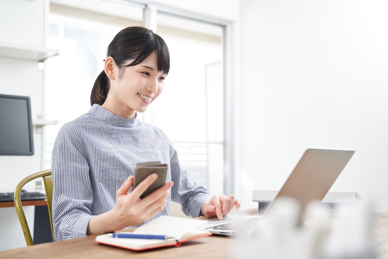 Woman at a desk working on a laptop with a cellphone in her hand