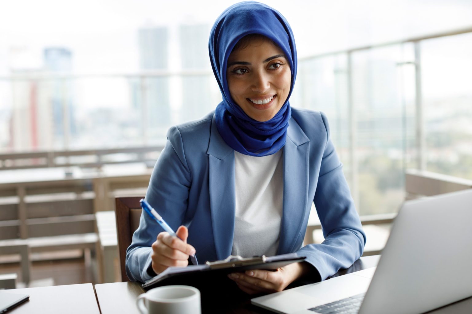 Woman working at a laptop writing on a clipboard and smiling.