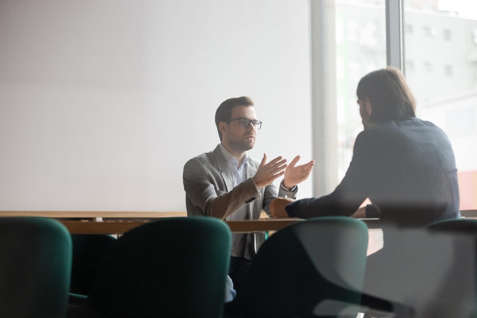 Two men sitting in a boardroom together and one is talking to the other with his hands explaining.