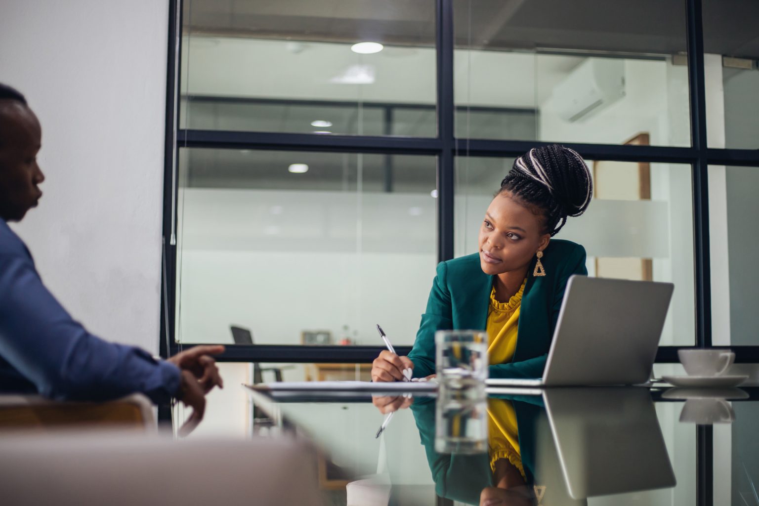 Woman sitting in a boardroom with an employee and writing notes with a laptop nearby.