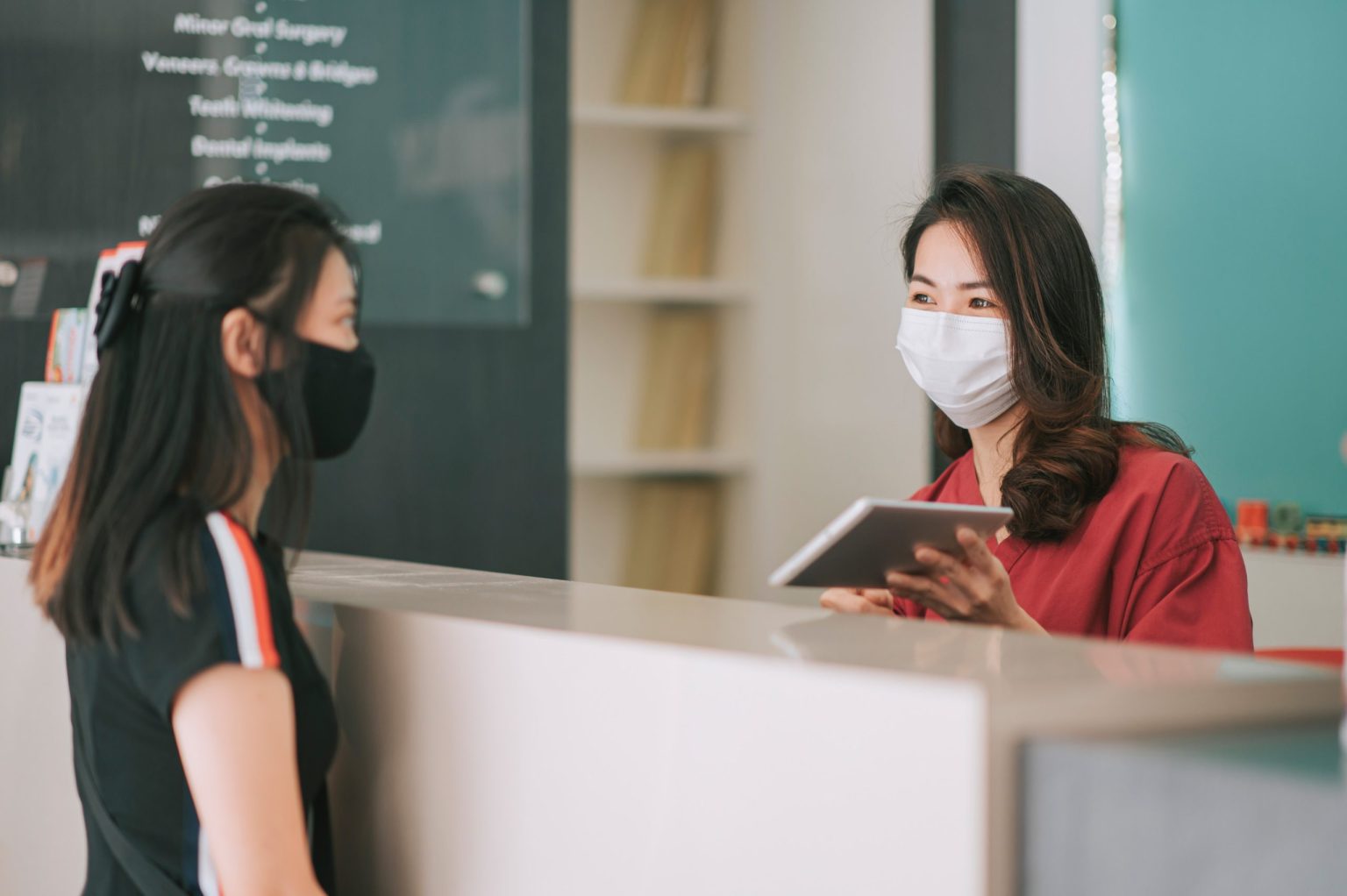 Two women with masks on at a dental clinic and the assistant has a tablet and is helping the patient.