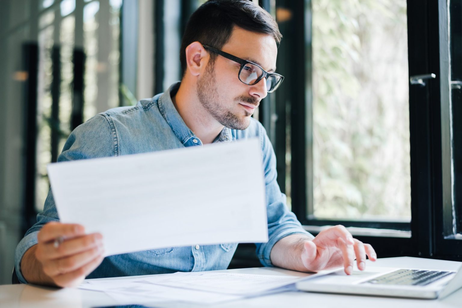 Man with glasses on working at a table in front of windows on a laptop with papers in his hands.