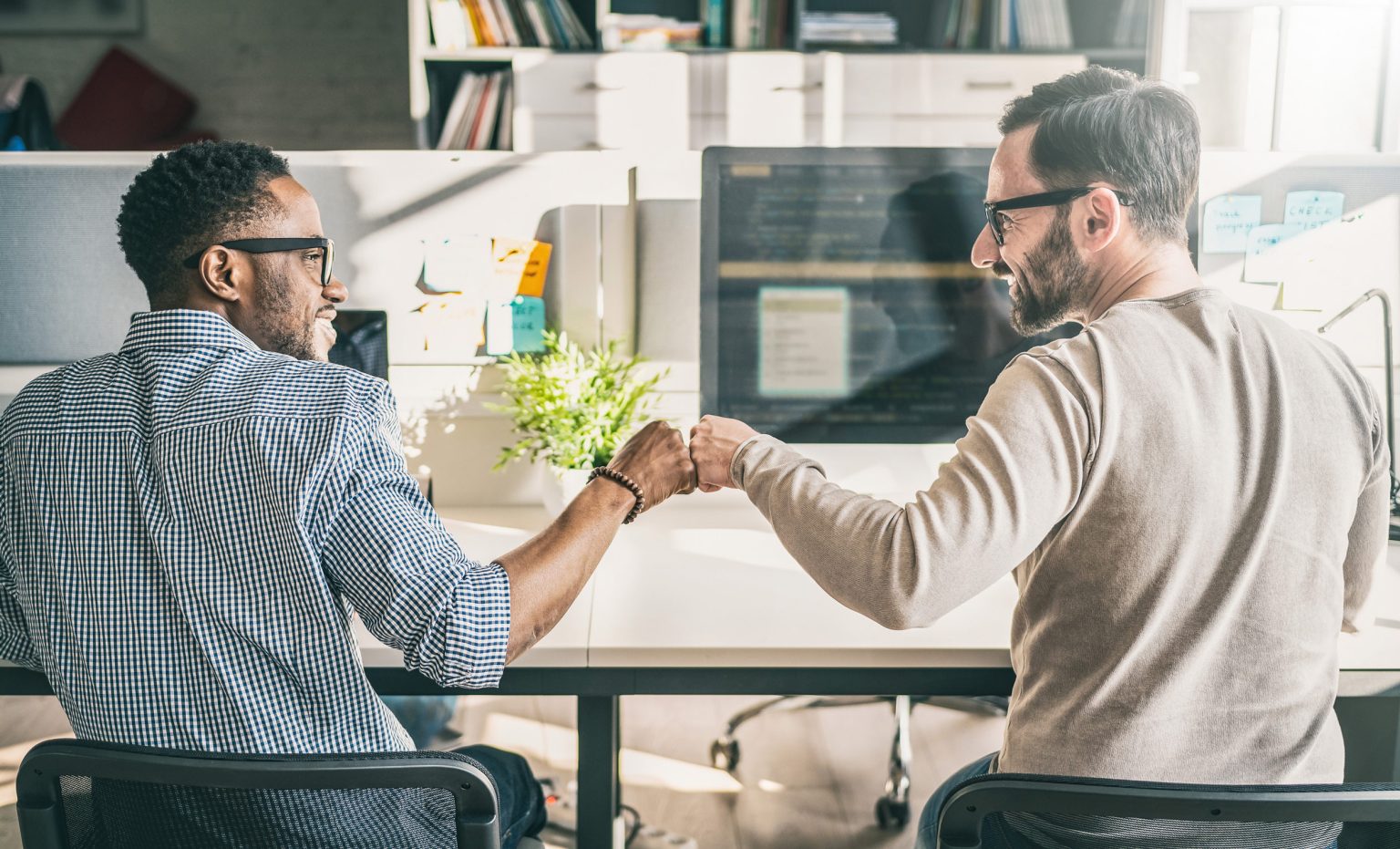Two male office workers at a desk looking at each other and doing a fist bump and smiling.