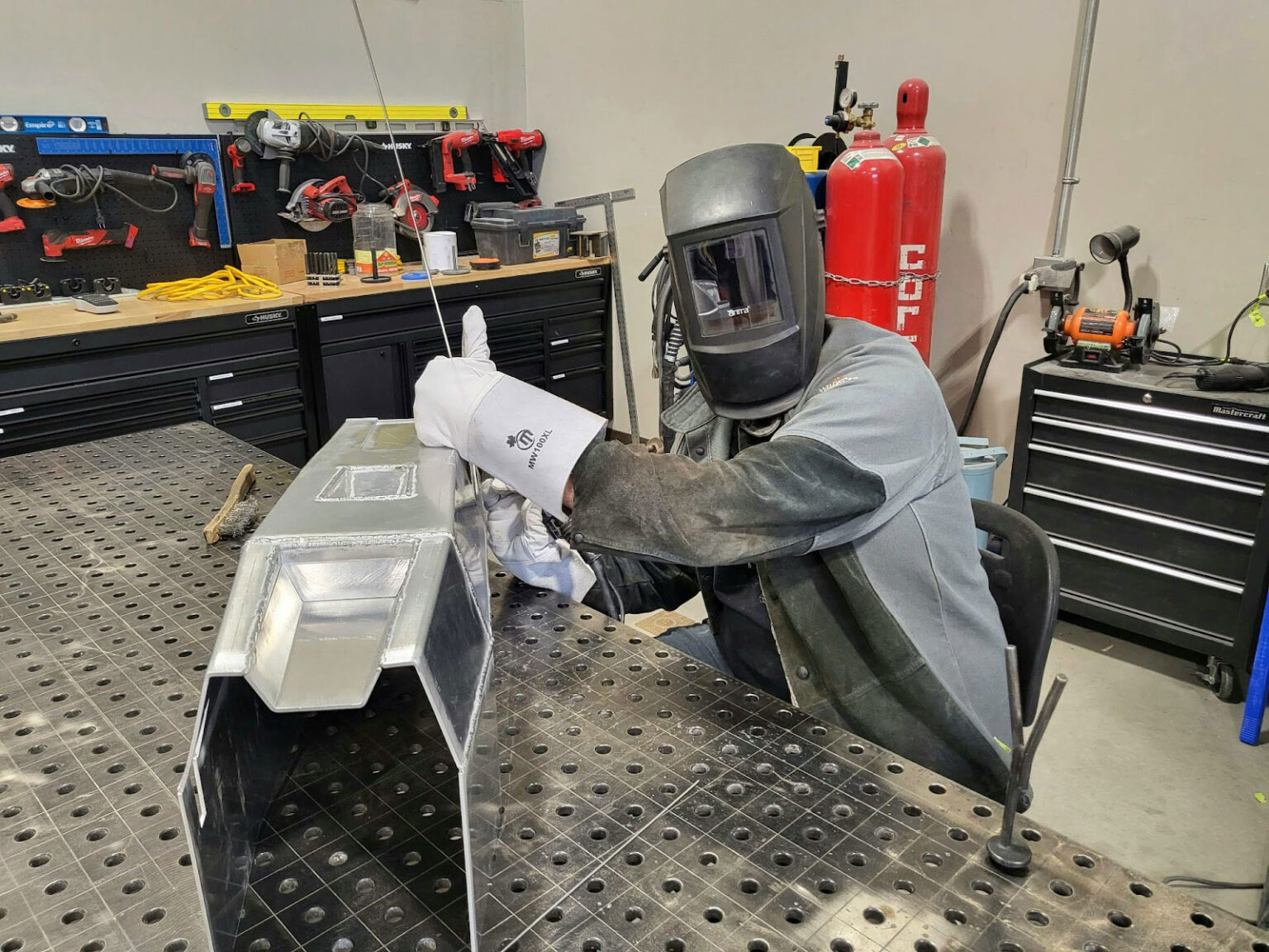 A person in welding gear cleans a metal object on a workbench, surrounded by the week's tools and equipment that fuel innovation in the bustling workshop.