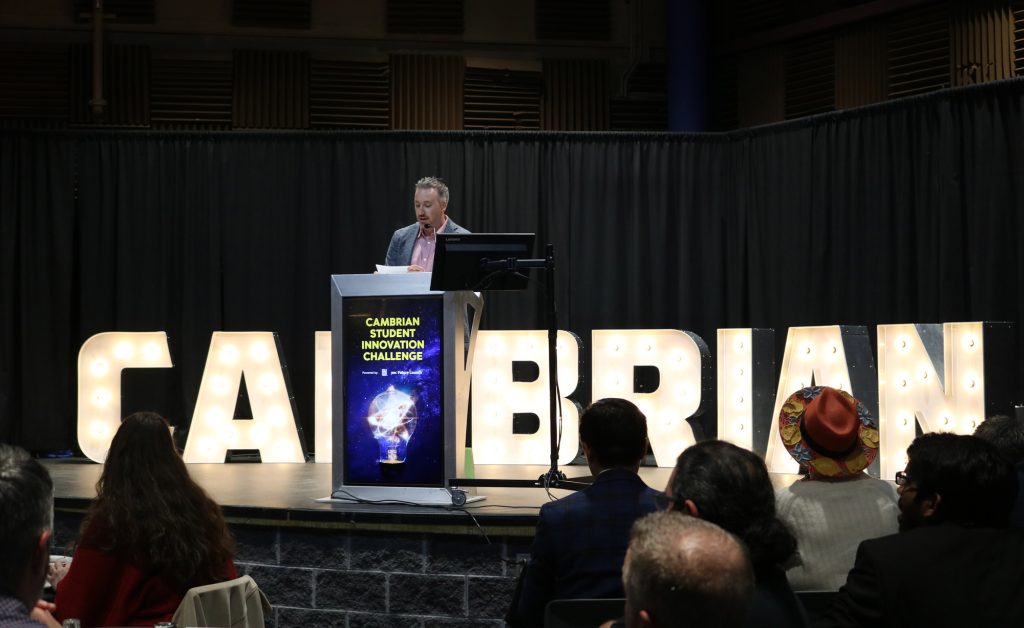 A speaker at a podium addresses an audience at the Cambrian Student Innovation Challenge event, with illuminated "CAMBRIAN" letters in the background, highlighting the spirit of innovation endorsed by RBC Future Launch.