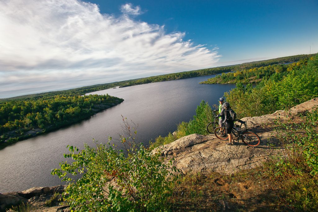 A picture of two individuals walking with their bikes and behind is Lake Laurentian