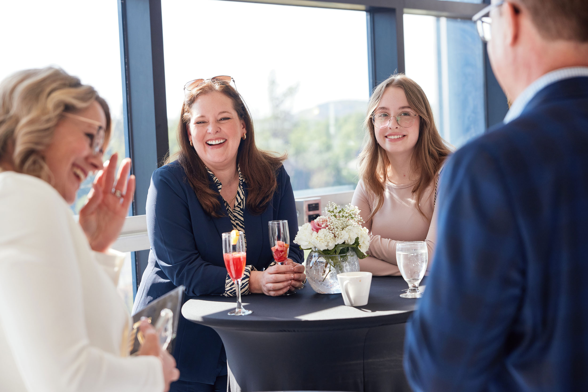 Four people stand around a high table with drinks and flowers, engaging in conversation and laughter.