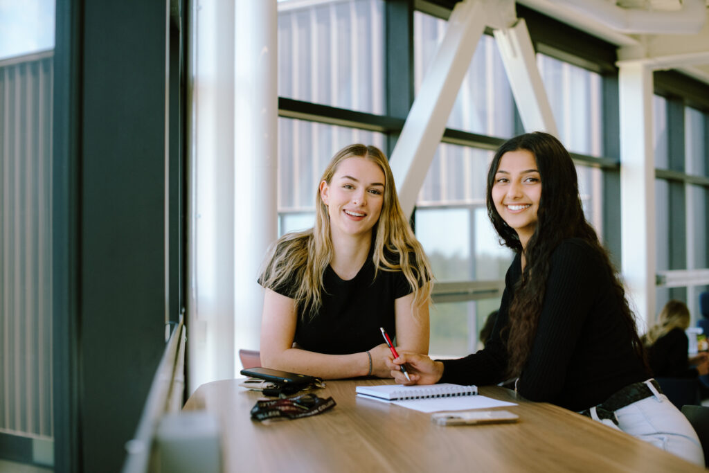 Two students sitting at a table on campus with a book and a pen in front of them.