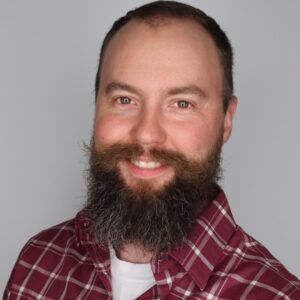 Spencer Julien, with a full beard and mustache, wearing a red plaid shirt over a white t-shirt, smiles at the camera against a plain light background.