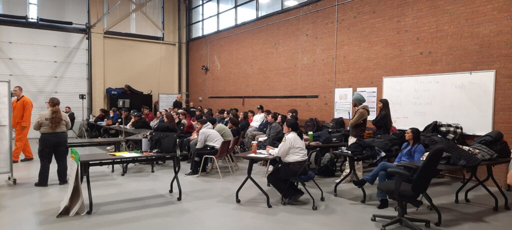 A large group of people sits in a room with a high brick wall at Cambrian College, listening to a presentation. Tables and chairs are arranged, notes are visible on whiteboards, and the Crisis Intervention Team offers insights.