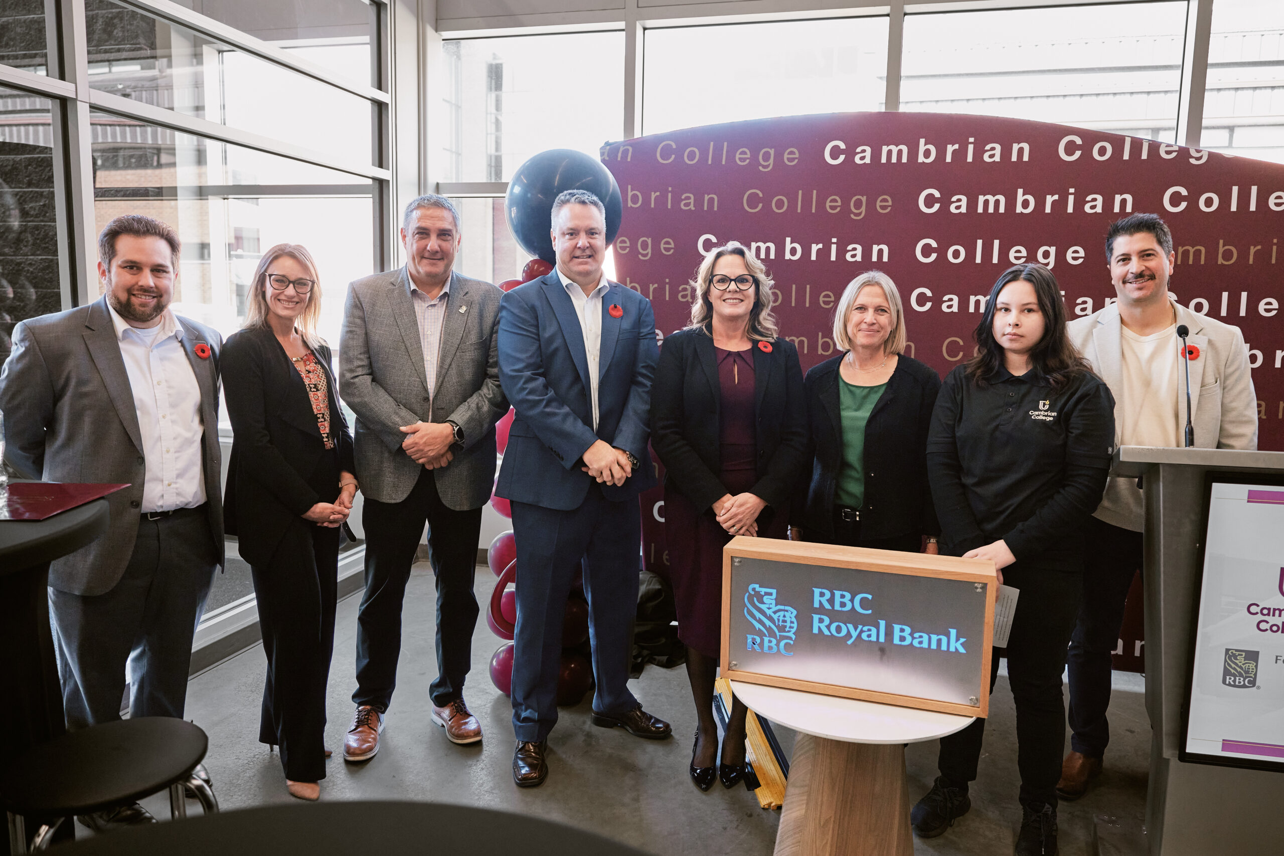A group of eight people stands in a row, some wearing poppies, with a Cambrian College backdrop and an RBC Royal Bank sign visible, highlighting the collaborative spirit fostered by the RBC Foundation.