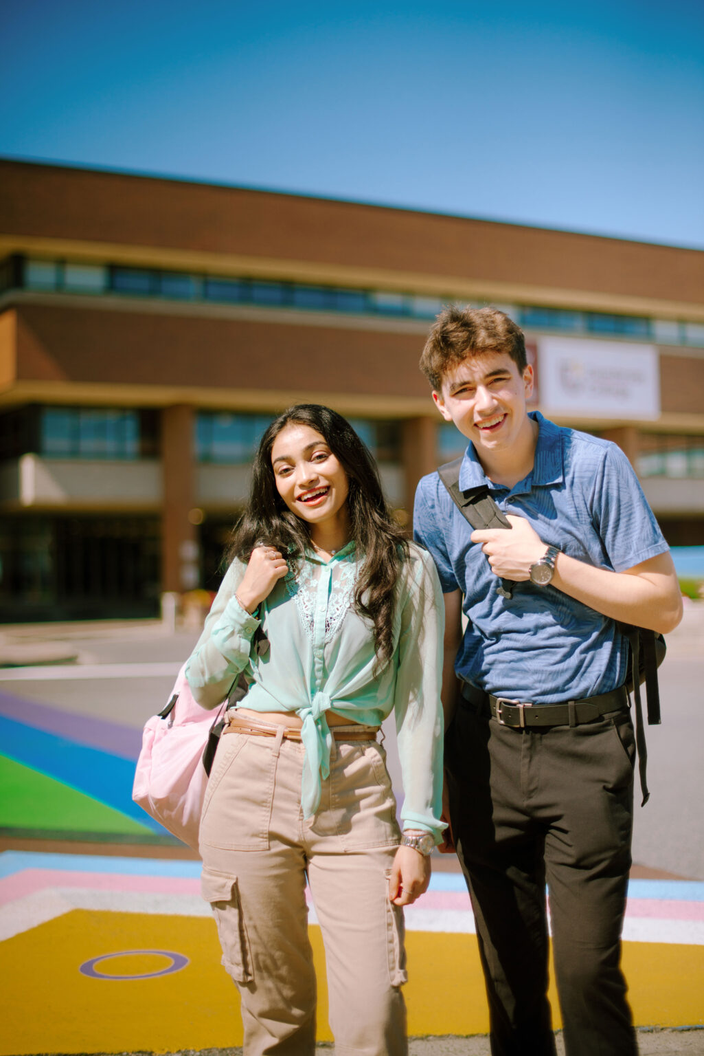 Two students standing outside a building, smiling, with backpacks over their shoulders.
