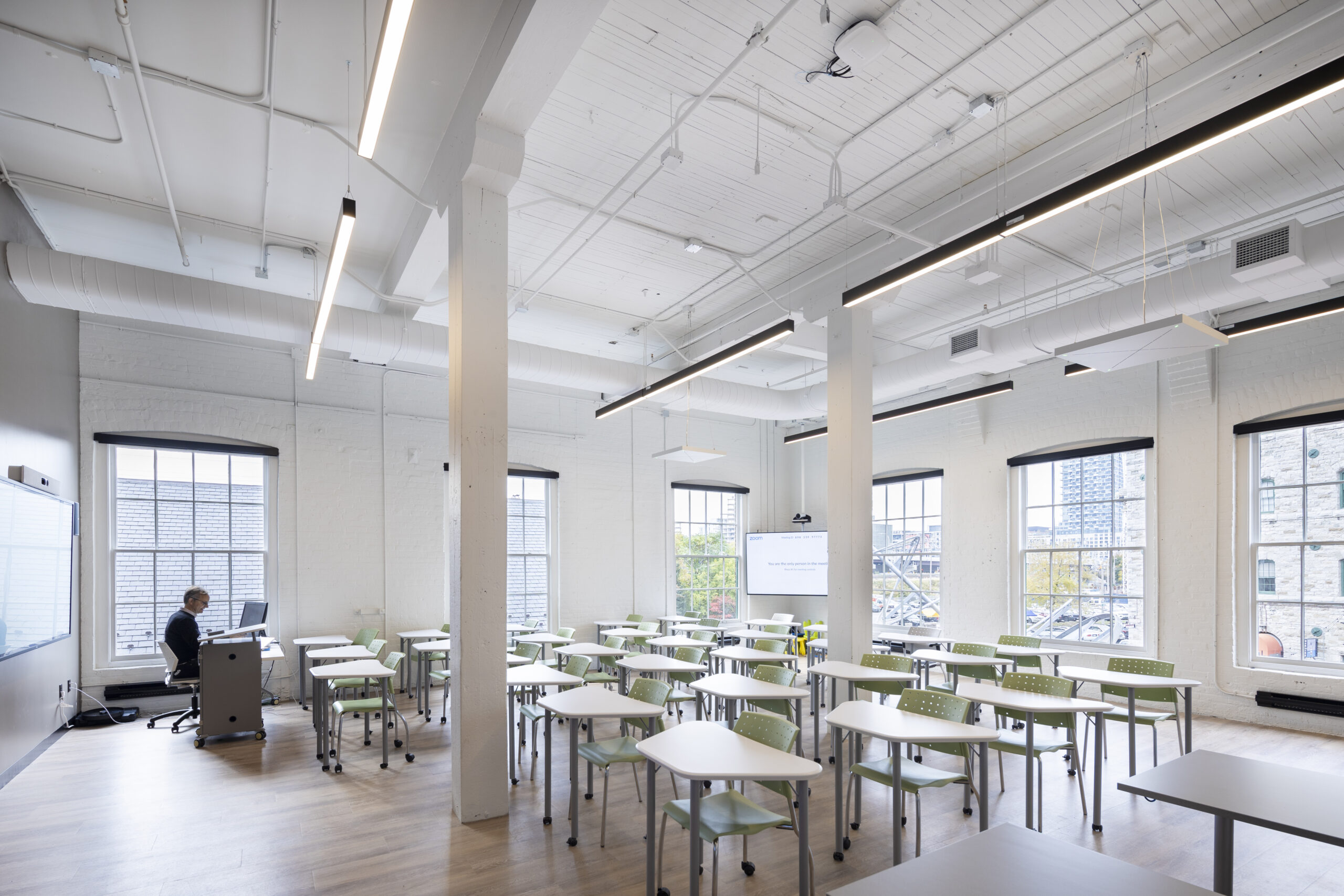 A modern, bright classroom with empty desks and chairs; one person sits at a teacher’s podium near large windows and a screen displaying a presentation.