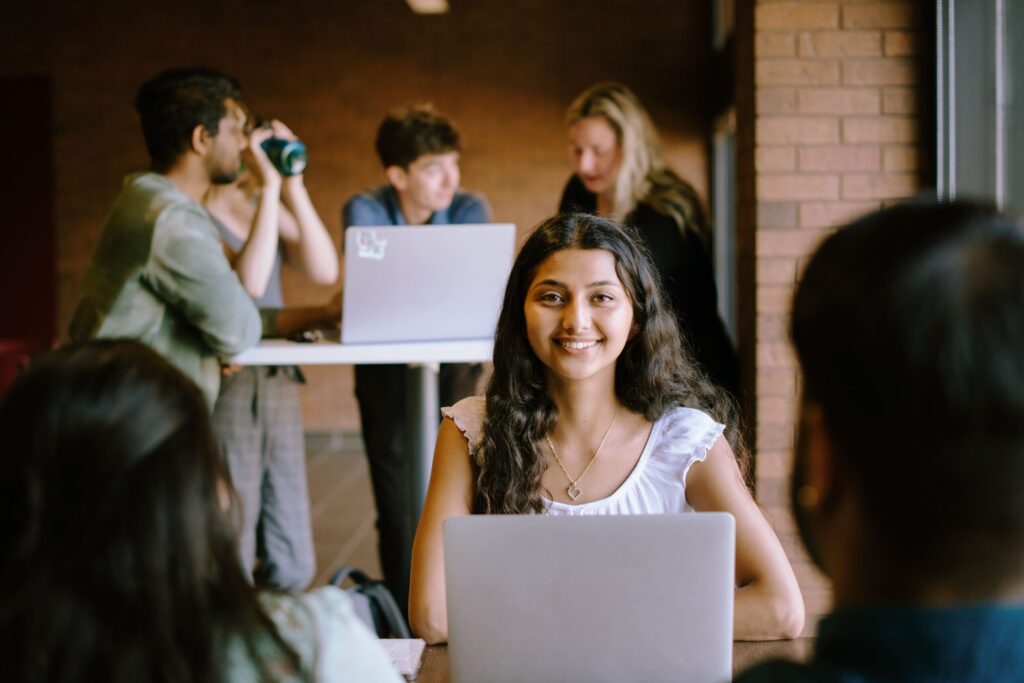 A student sits at a table with a laptop, smiling at the camera. In the background, four people interact around another table with laptops.