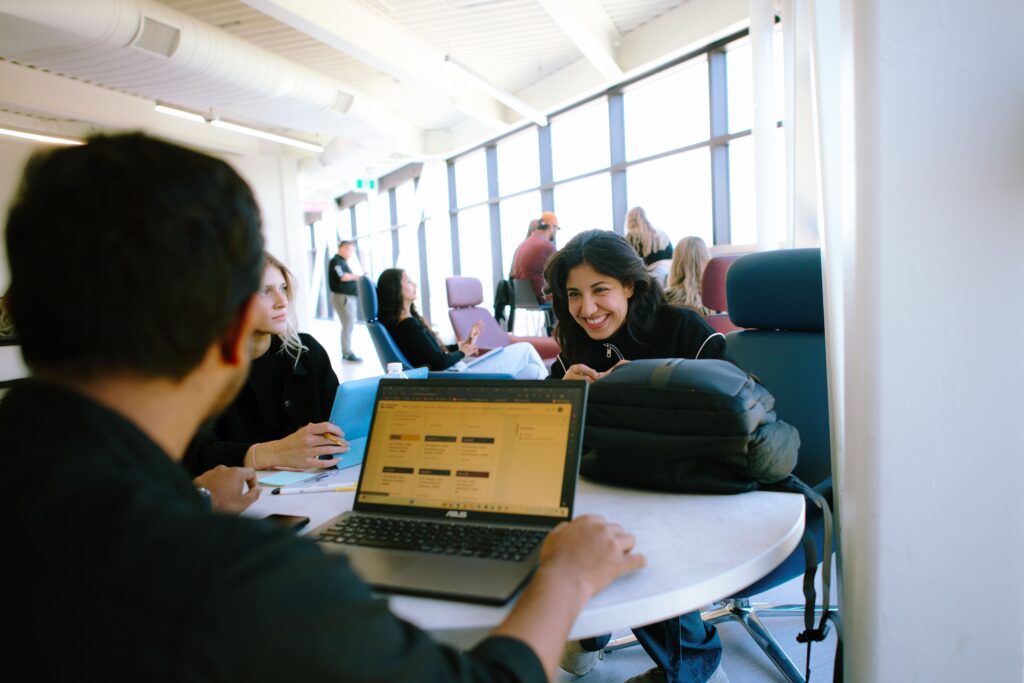 Three people are sitting at a round table in a bright room; one is using a laptop, while the others are talking and smiling. Other people are visible in the background.