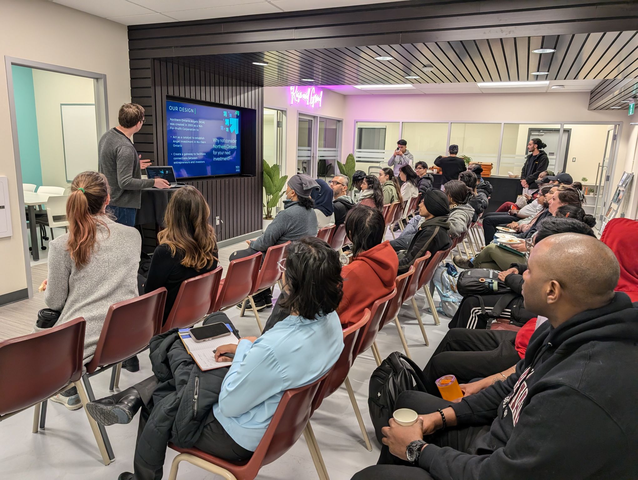 A group of people sit in rows facing a screen where a presenter gives a talk in the modern, well-lit Hanson Venture Lab.