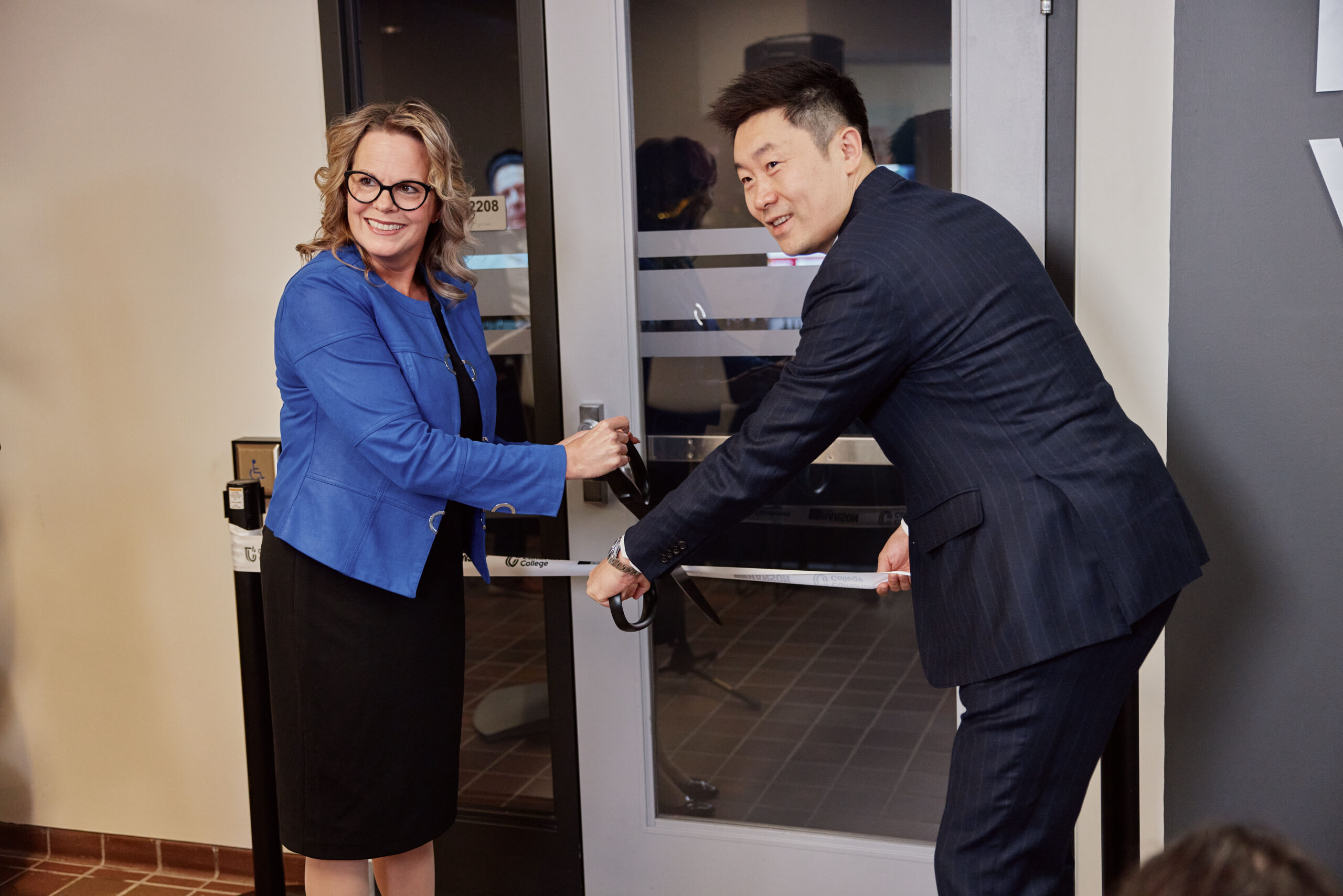 Two people in business attire cut a ribbon together at a doorway, likely marking the opening of a new office or facility.