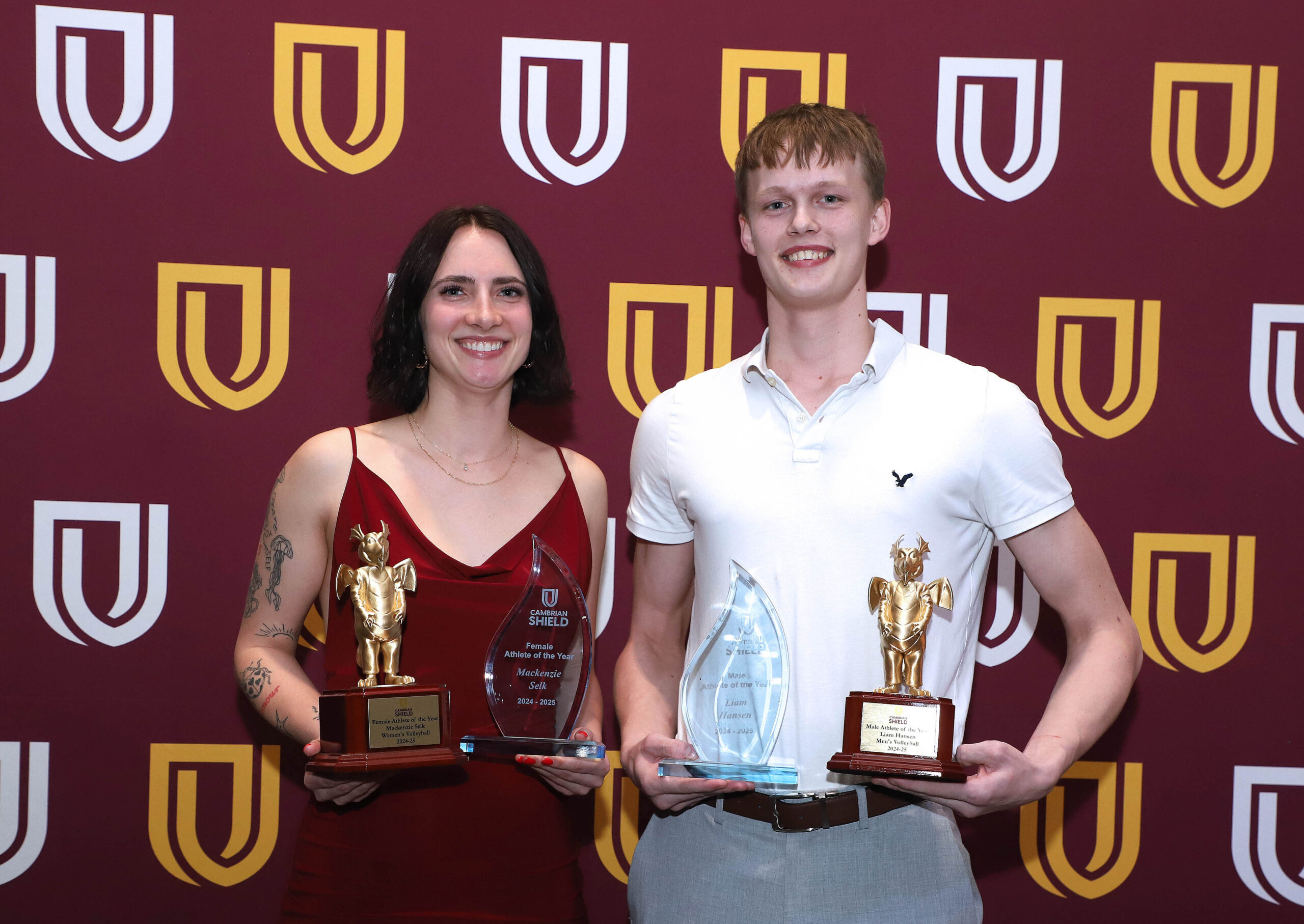 Two people stand in front of a maroon backdrop with yellow and white logos, each holding two trophies and smiling at the camera.
