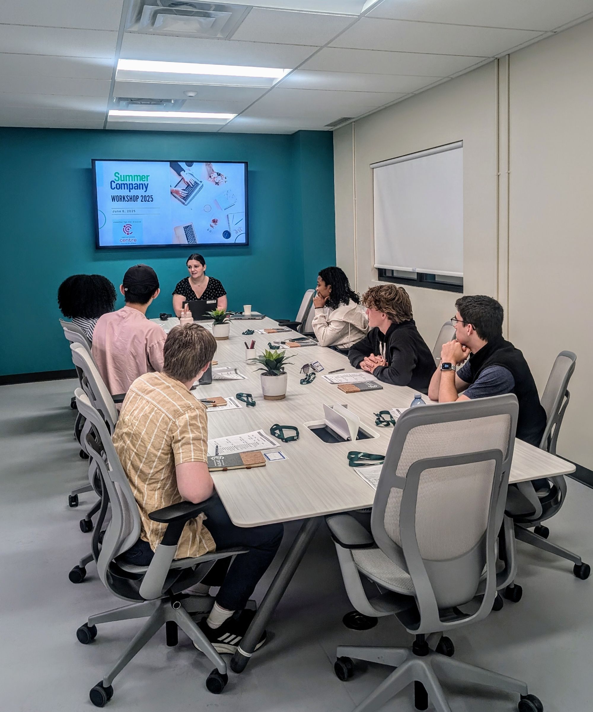 Seven people sit around a conference table in a meeting room, listening to a woman speaking. A screen displays "Summer Company Workshop 2023," hosted by the Hanson Venture Lab.