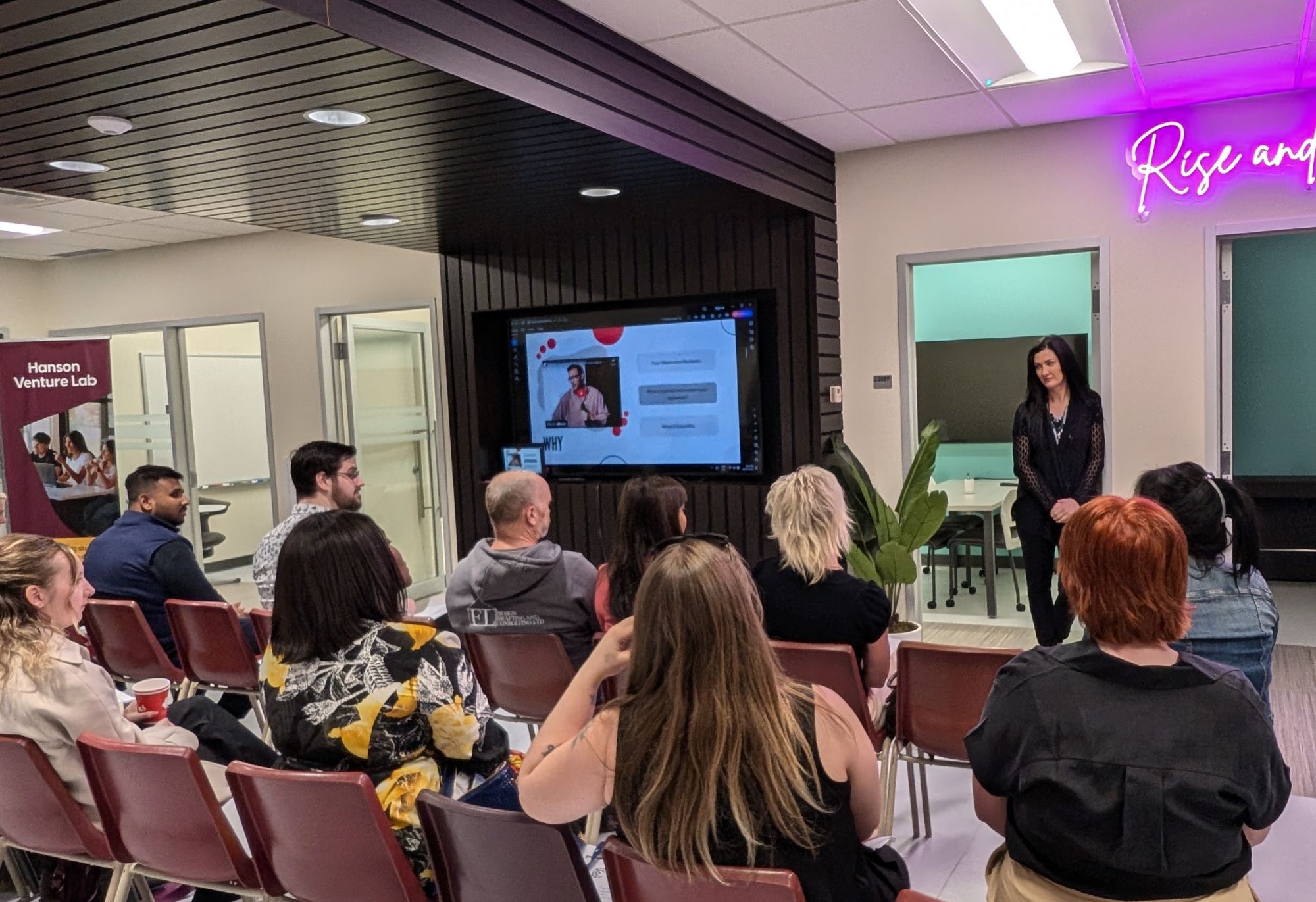 A woman stands at the front of a room speaking to a seated audience; behind her, a screen displays a Hanson Venture Lab video call presentation.