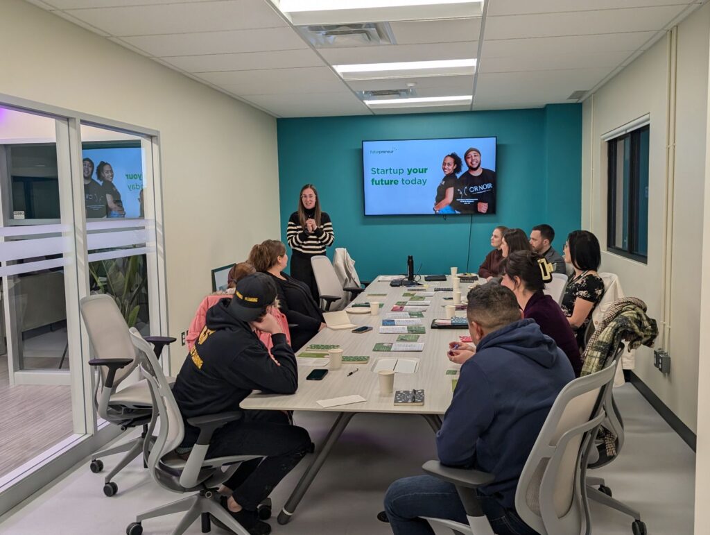 A presenter stands and presents to a group of people seated around a conference table in the Hanson Venture Lab, with a screen displaying “Startup your future today.”.