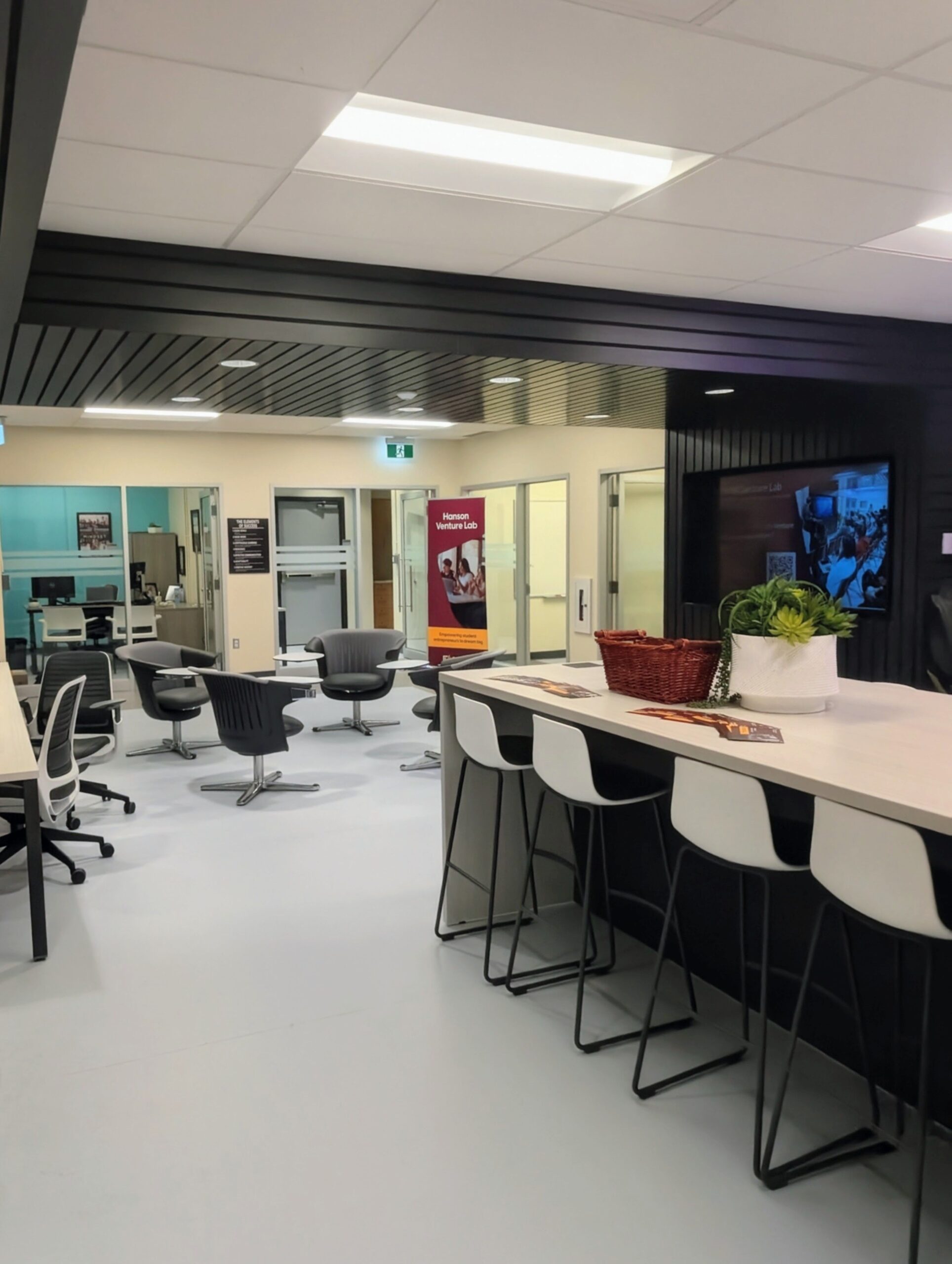 Modern office space with high table and stools in the foreground, round tables and chairs in the background, and glass-walled meeting rooms visible—ideal for collaboration at the Hanson Venture Lab.